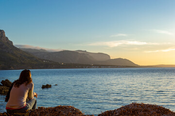 A young and beautiful girl dressed in casual clothes looking at the sunset at a beautiful beach in Artà Mallorca Balearic Islands Spain with a impressive view of the sea and the Tramuntana mountains