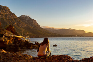A young and beautiful girl dressed in casual clothes looking at the sunset at a beautiful beach in Artà Mallorca Balearic Islands Spain with a impressive view of the sea and the Tramuntana mountains