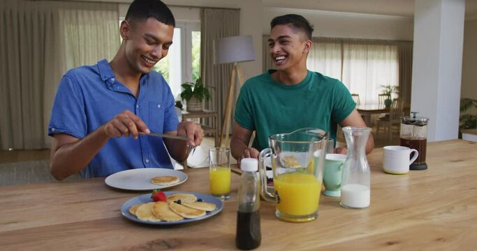 Happy mixed race gay male couple having pancakes for breakfast and laughing in kitchen