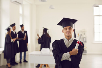 Confident and proud student in the form of a university graduate and with a diploma in hand stands...