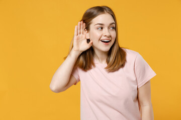 Young caucasian curious smiling woman 20s with nude make up in casual basic pastel pink t-shirt trying to hear you overhear listening intently isolated on yellow color background studio portrait