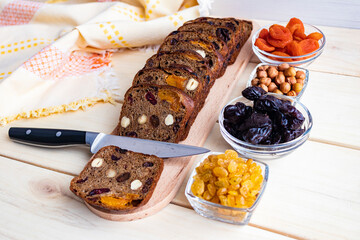 Freshly baked rye bread with dried fruits, cut on a board, a knife and raisins, dried apricots, prunes and hazelnuts on a wooden background, close-up.