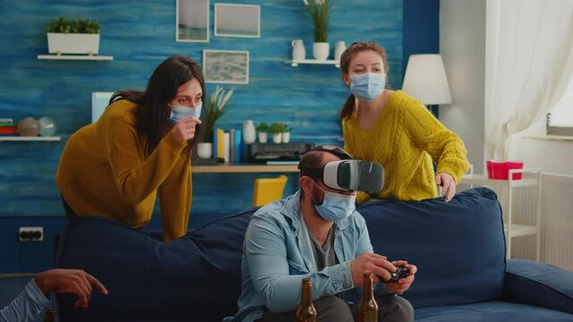 Excited man spending time with friends experiencing playing games with vr headset wearing face mask to prevent coronavirus spread in global pandemic. Diverse people having fun at new normal party