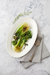 Top View of Charred Baby Bok Choy on a White Oval Plate on a Marble Counter; Gray Napkin and Fork Beside Plate