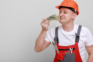 Happy worker in red uniform, protective hard hat holding bundle of dollars, sniffing of cash money on white background. Male worker for advertisement.