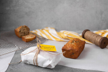 One Loaf of Banana Nut Bread Wrapped in Parchment Paper with Yellow Tag; One Loaf Unwrapped and Aother Sliced in Background on Cooling Rack