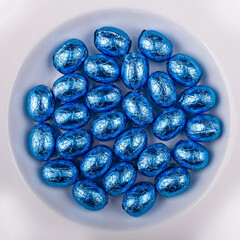 Top view of a pile of blue, wrapped in shiny tinfoil, chocolate Easter eggs in a white round bowl