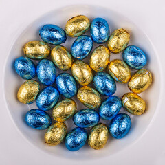 Top view of a pile of blue and Yellow golden, wrapped in shiny tinfoil, chocolate Easter eggs in a white round bowl