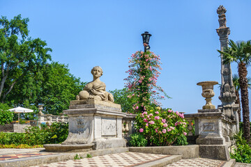 Fototapeta na wymiar Panorama of old park decorations in Massandra palace. There are sphinx with female head, bowl & column in baroque style & lots of flowers. Shot near Yalta, Crimea
