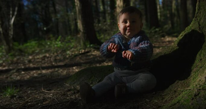 A little baby is sitting on the ground in the woods and is playing with a stick on a sunny spring day