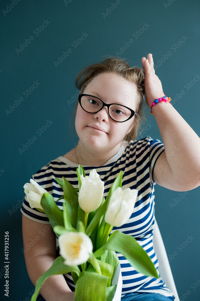 Wall mural Portrait of  teenage girl with Down syndrome standing with flowers on  blue background. Disability children. World Down syndrome day.