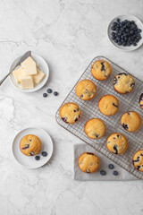 Top View of Homemade Blueberry Muffins on a Cooling Rack; One on a White Plate; One on a Gray Napking; Butter on Plate; Blueberries on Plate; Gray and White Marble Countertop