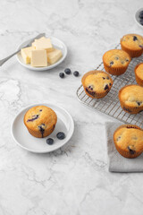 Homemade Blueberry Muffins on a Cooling Rack; One on a White Plate; One on a Gray Napking; Butter on Plate; Blueberries on Plate; Gray and White Marble Countertop