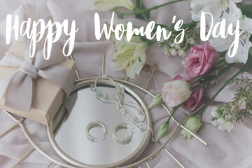 Happy womens day text on stylish gift box, modern jewellery and spring bouquet on boho mirror on soft fabric. Modern greeting card. Stylish handwritten sign. International womens day