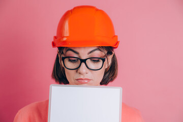 Pretty thoughtful  woman worker builder hold white sign board blank against pink background. Building helmet.
