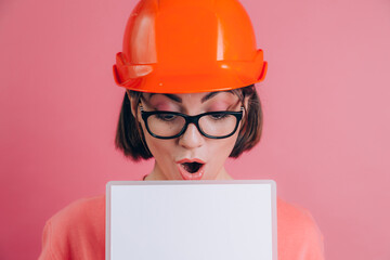 Pretty amazed surprised woman worker builder hold white sign board blank against pink background. Building helmet.