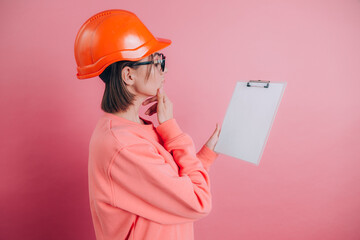 Pretty thoughtful  woman worker builder hold white sign board blank against pink background. Building helmet.