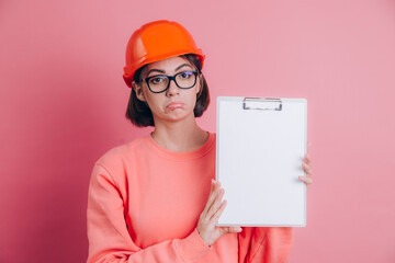 Sad unhappy disappointed woman worker builder hold white sign board blank against pink background. Building helmet.