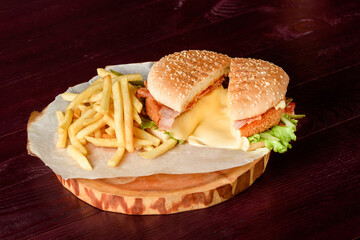 Burger in a cut with cutlet, melted cheese, bacon, lettuce, tomatoes and potatoes on parchment on a wooden platter on a wooden background.