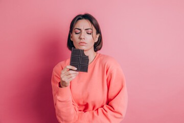 Beautiful sad woman with abdominal pain with chocolate bar on pink background and bright makeup