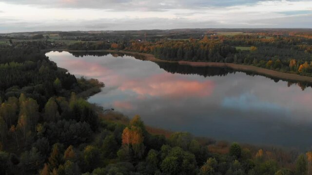 Jesienny widok na jezioro pośr&oacute;d drzew o zachodzie słońca. W wodzie widoczne odbicie chmur. Ujęcie z drona, dolly out, 4K. 