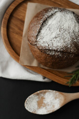 Round black bread on baking paper and a wooden spoon with flour on a black background. Close-up.