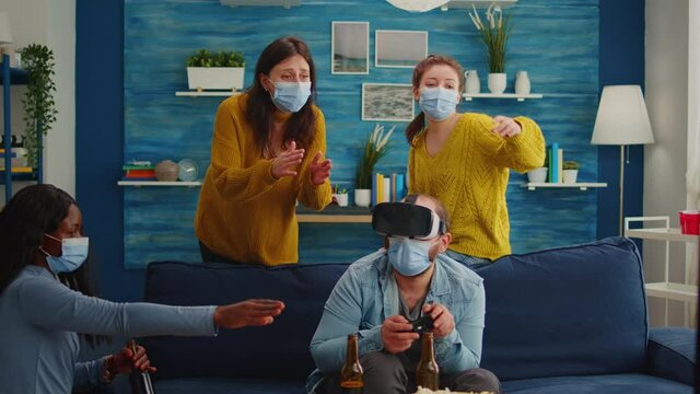 Group of multi ethnic friends with face mask enjoying playing video games using vr headset goggles in living room keeping social distancing agaist covid19. Diverse people having fun at new normal