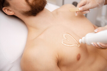 Laser depilation concept for men. Beauty salon worker applies gel to guy body before hair removal