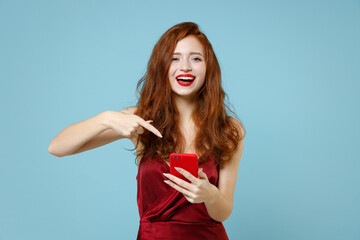 Young caucasian excited readhead curly woman in red party evening dress gown point index finger on mobile cell phone read hot news surfing internet isolated on pastel blue background studio portrait.