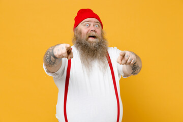 Fototapeta na wymiar Fat pudge obese chubby overweight tattooed bearded surprised fun shocked man has big belly in white t-shirt red hat suspenders point index finger camera on you isolated on yellow background studio