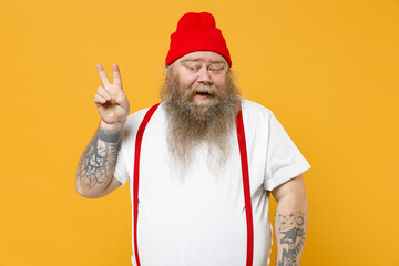 Fototapeta na wymiar Fat pudge obese chubby overweight tattooed bearded friendly kind man 30s has big belly in white t-shirt red hat suspenders show victory v-sign gesture isolated on yellow background studio portrait.
