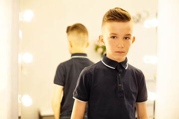 10-year-old boy in a hairdresser after a fashionable haircut in front of a mirror. A small child in a beauty salon looks at the camera posing. Hairdresser rape service.