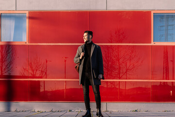 Young businessman with backpack and laptop in front of a red wall at sunset looking at the sun