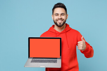 Young caucasian smiling web designer bearded man 20s in casual red orange hoodie hold laptop pc computer, blank screen workspace area show thumb up gesture isolated on blue background studio portrait.