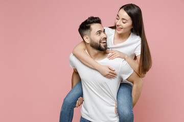 Naklejka premium Young cheerful lovely couple two friends man woman in white basic blank t-shirts giving piggyback ride to joyful sits on back fooling around isolated on pastel pink color background studio portrait
