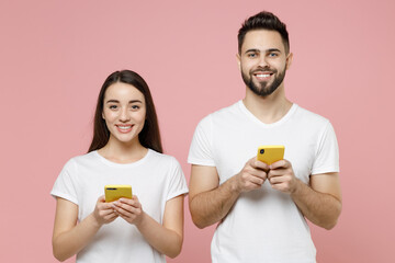 Young couple two friends man brunette woman 20s in white basic blank print design t-shirts hold in hand using mobile cell phone chatting typing sms isolated on pastel pink background studio portrait.