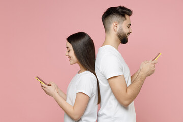 Young couple two friends man woman 20s in white blank design t-shirts hold in hand using mobile cell phone chatting typing sms standing back to back isolated on pastel pink background studio portrait.