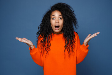 Shocked amazed surprised young african american woman wearing casual basic bright orange sweatshirt standing keeping mouth open spreading hands say wow isolated on blue background studio portrait.