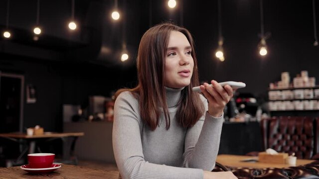 Smiling young woman recording voice message on smartphone. Beautiful girl using virtual assistant app sitting at coffee shop. Voice recognition, audio messages, using translator on mobile phone