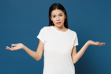 Young confused mistaken disappointed mexican latin woman 20s wearing white casual basic t-shirt spreading hands oops gesture looking camera isolated on dark blue color background studio portrait.