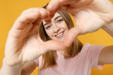 Young lovely romantic woman 20s with nude make up wearing casual basic pastel pink t-shirt look through close up shape heart with hands, heart-shape sign isolated on yellow background studio portrait