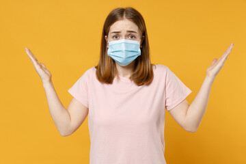 Young indignant confused woman 20s in basic pastel pink t-shirt in sterile face mask from coronavirus virus covid-19, pandemic quarantine spread hands isolated on yellow background studio portrait.