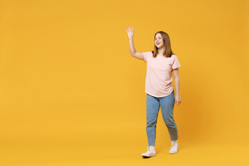Fototapeta na wymiar Full length of young friendly smiling student woman 20s with nude make up in casual basic pastel pink t-shirt, jeans waving hand greeting someone say hi isolated on yellow background studio portrait.