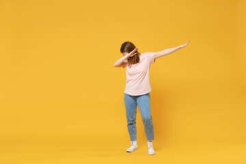 Full length of young woman in casual basic pastel pink t-shirt, jeans doing dab hip hop dance hands move gesture youth sign hiding covering face isolated on yellow color background studio portrait