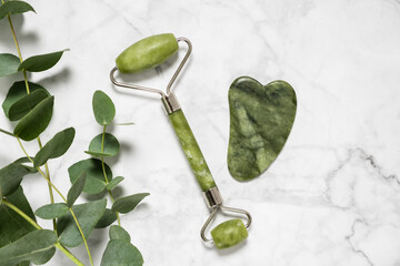 Green jade roller and gua sha stone for facial massage and eucalyptus branch on marble background....