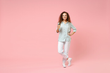 Full length young black african fun happy smiling stylish curly student woman 20s in blue shirt holding paper cup of coffe arm akimbo on waist isolated on pastel pink color background studio portrait.
