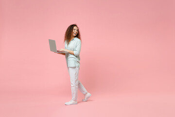 Full length young black african fun happy smiling curly student woman 20s wearing blue shirt hold laptop pc computer walking looking aside isolated on pastel pink color background studio portrait.