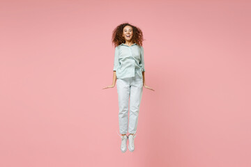 Full length young black african fun happy surprised overjoyed joyful happy smiling energetic excited woman 20s wearing blue shirt jumping high isolated on pastel pink color background studio portrait.