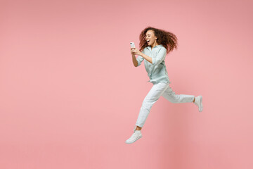 Fototapeta na wymiar Full length side view young black african fun happy smiling curly student woman 20s in blue shirt holding mobile cell phone chatting jump high run isolated on pastel pink background studio portrait.