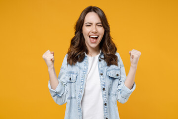 Young excited overjoyed happy fun brunette expressive woman in stylish casual denim shirt white t-shirt do winner gesture clench fist celebrate isolated on yellow color background studio portrait.